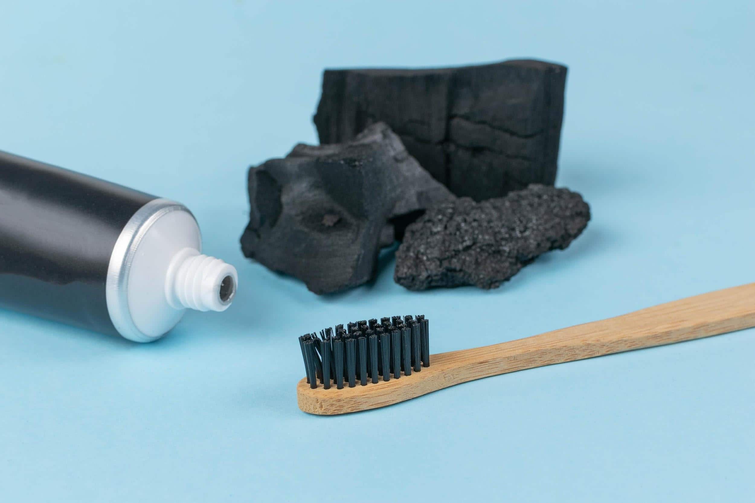 Wooden toothbrush with black charcoal toothpaste, tube of black toothpaste and chunks of activated charcoal on a light blue background